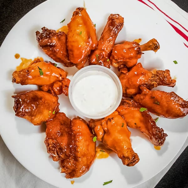 A white plate of saucy chicken wings arranged around a small cup of white dipping sauce.