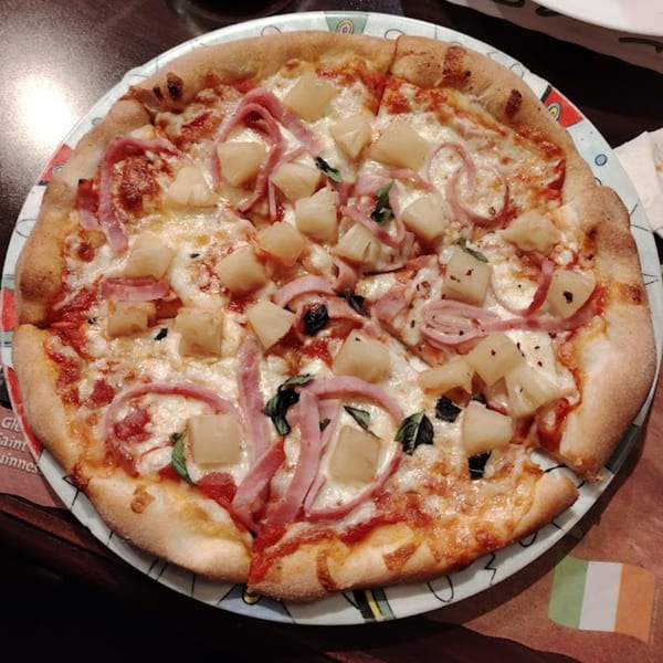 A Hawaiian pizza from Touch of Italy with pineapple, ham, and basil on a colorful plate.